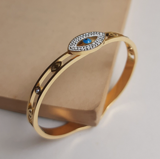 A gold-plated bracelet with a central evil eye design surrounded by crystals, displayed against a beige background.